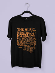 Black t-shirt with orange text and piano graphic on a hanger against a light purple background